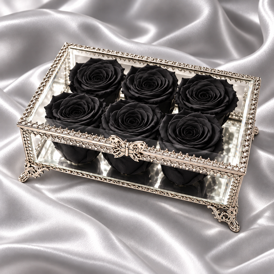 A rectangular silver-toned decorative box with a mirrored lid and six black preserved roses arranged inside, set against draped silver satin fabric.