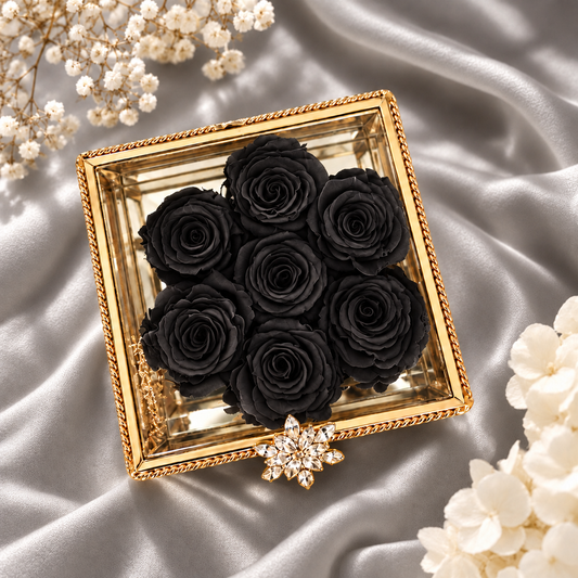 A square gold-toned decorative box filled with seven preserved black roses, styled on draped satin fabric with white floral accents in the corners.