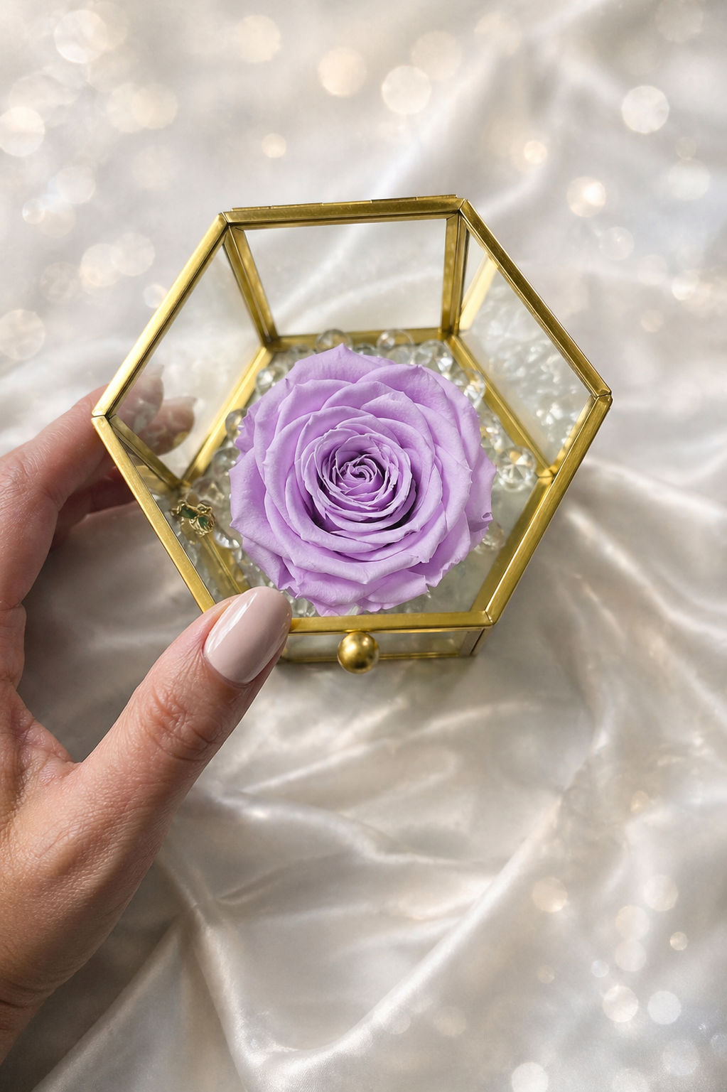 Hexagonal gold glass box with a preserved purple rose held by a hand on a soft white background