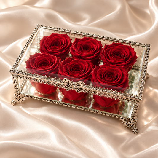 A rectangular glass keepsake box with ornate silver metal trim and feet, containing six preserved deep red roses, displayed on a draped champagne satin fabric.