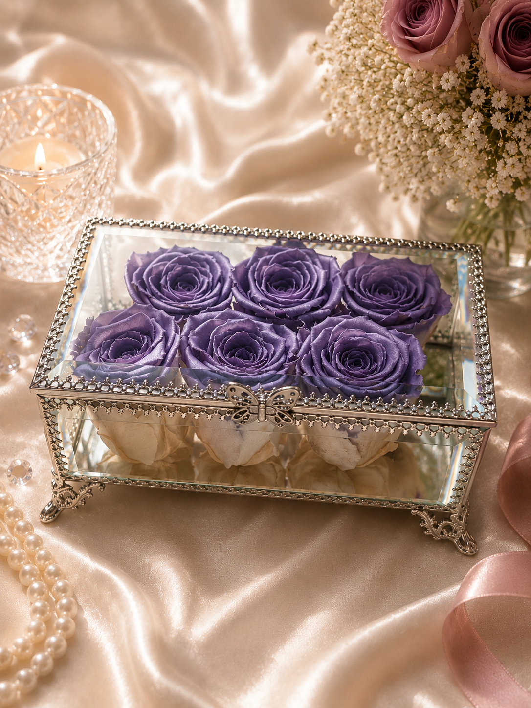 Decorative box with preserved purple and white roses on a satin surface with candles and flowers.