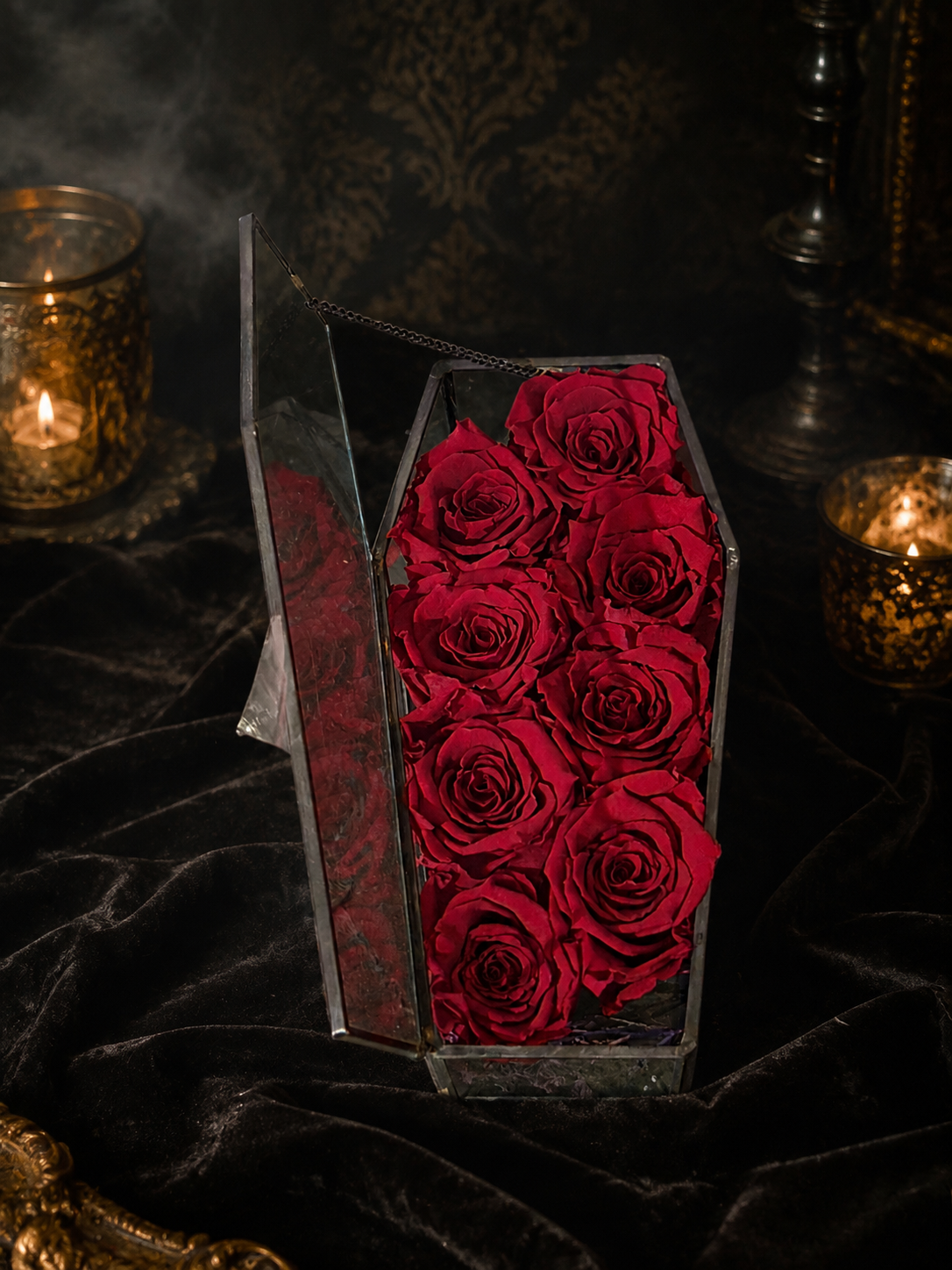 A coffin-shaped clear glass box filled with preserved deep red roses, styled on a dark velvet surface with lit candles and a smoky dark background.